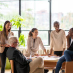 A small group of business professionals casually stand and sit around a boardroom table as they meet together to discuss company forecasts. They are each dressed professionally and have files in their hands as they meet together in the city.